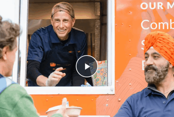 A smiling volunteer in a blue uniform hands food to a person with a backpack at a food truck with an orange background.