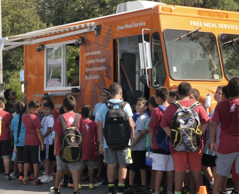 A group of children gather around an orange mobile food truck offering free meals, with some adults supervising.