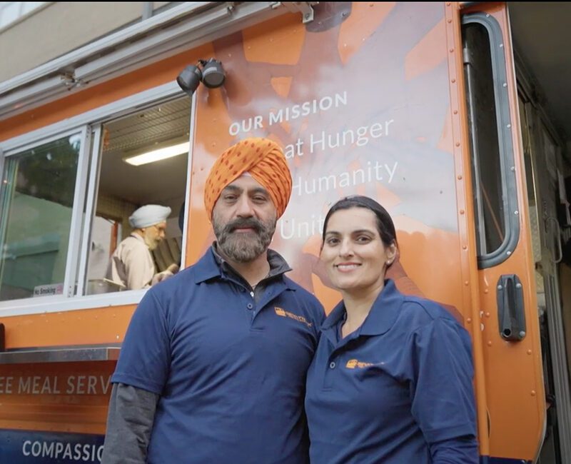 Two volunteers stand in front of a food truck with a mission statement, smiling and wearing matching blue shirts.