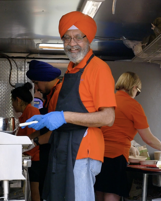 A smiling man wearing an orange turban, glasses, and a black apron prepares food in a busy kitchen with others in orange