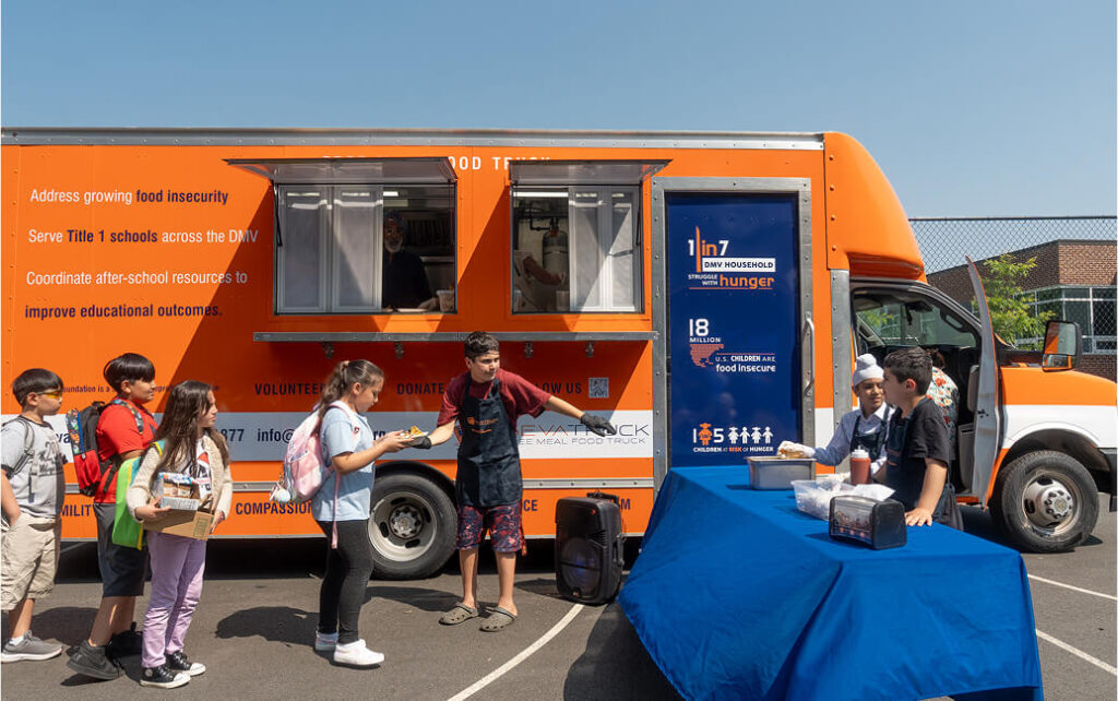 Children line up to receive food from a mobile food truck with volunteers serving in front of it.