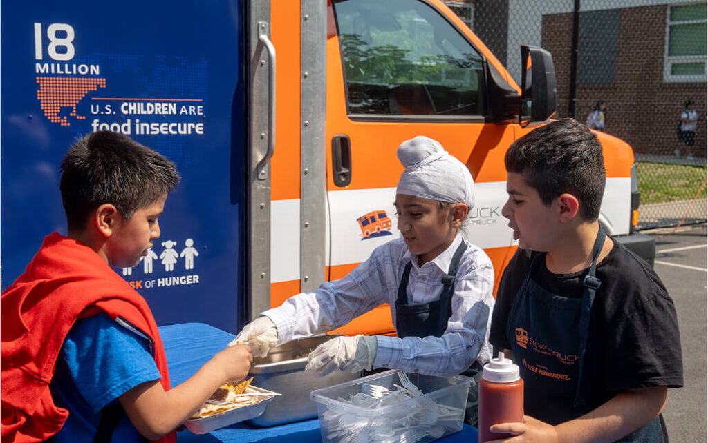 A girl in a headscarf and apron hands food to a boy in a blue shirt, with another boy holding a ketchup bottle nearby, in
