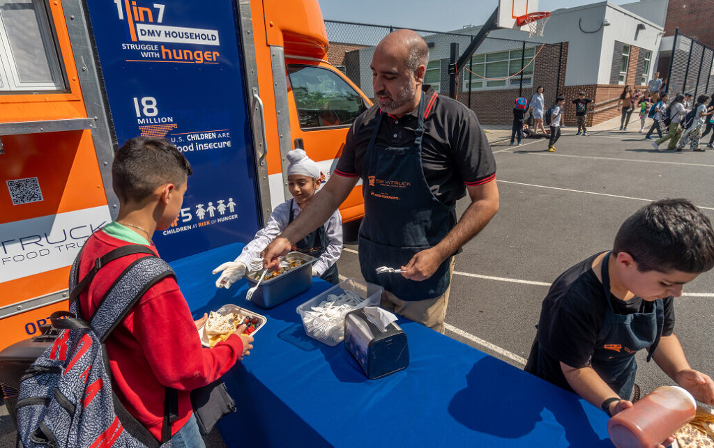A man serving food to children at a school event next to a colorful food truck with a basketball court in the background.