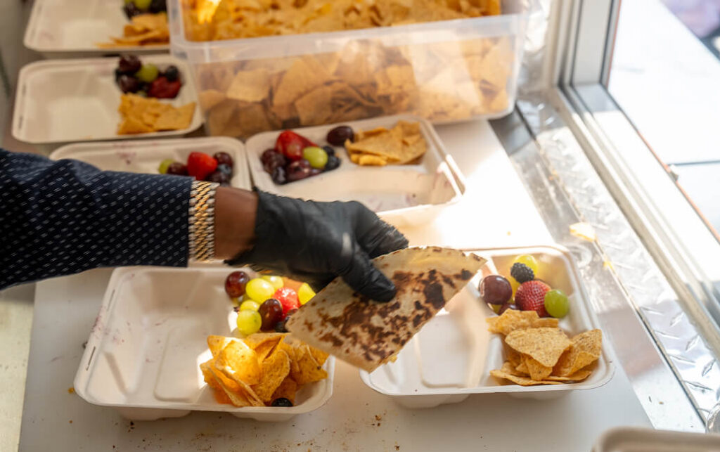 A person wearing a black glove and a polka dot shirt is placing a toasted quesadilla with grilled marks onto a tray of fruit