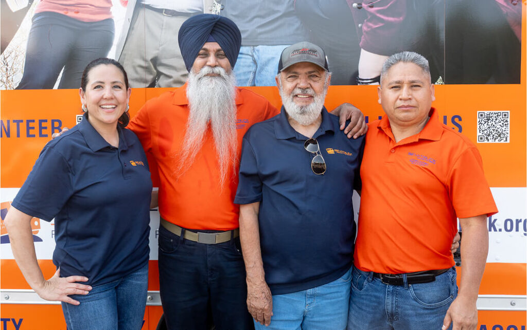 Four diverse volunteers stand together smiling in front of an orange banner, wearing navy and orange shirts.