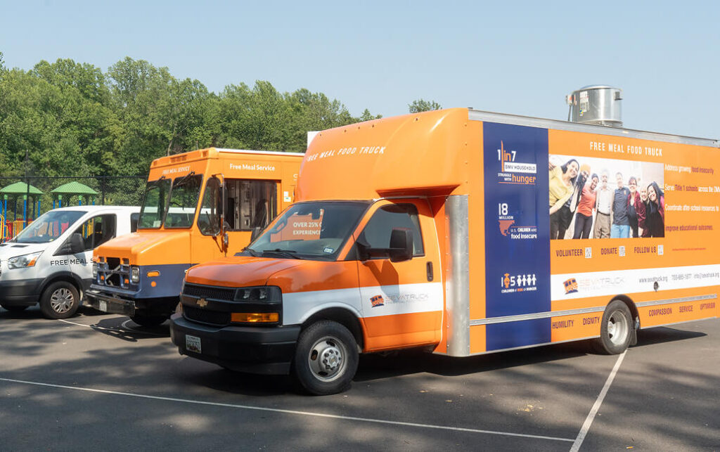 An orange and white mobile food truck parked alongside other vehicles, promoting free meals and community service.
