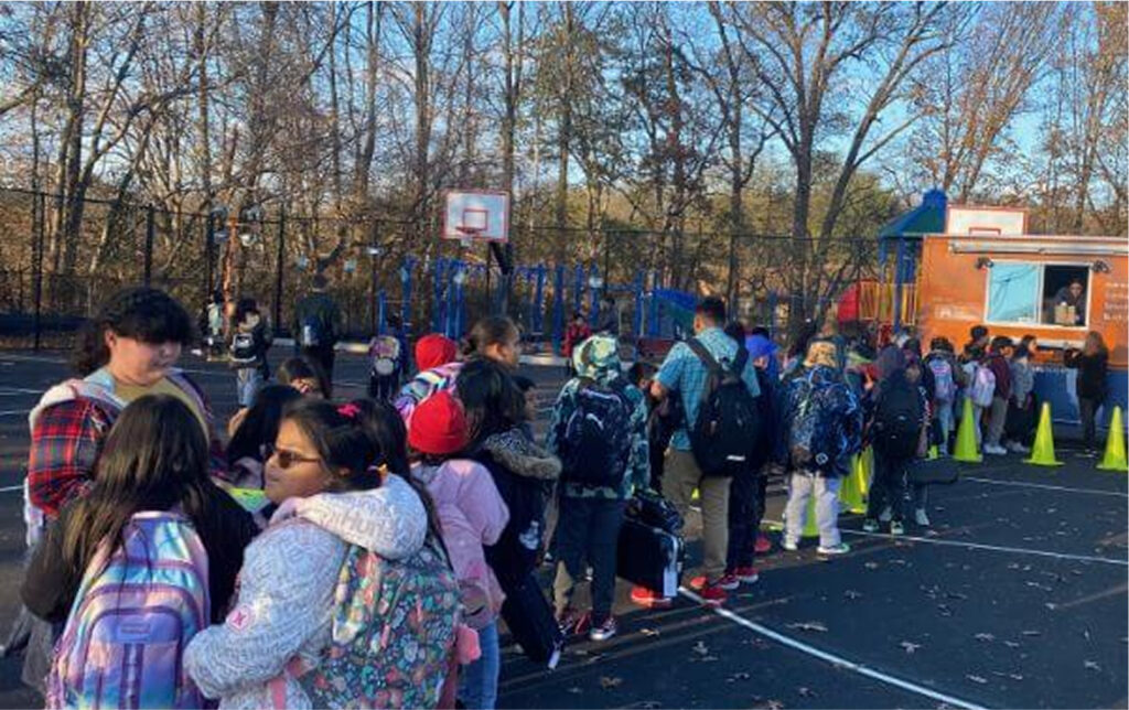 Children and adults line up outside a school gymnasium, with a playground and trees in the background on a clear day.