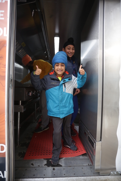 Two children in winter jackets and hats give thumbs up inside a food truck.