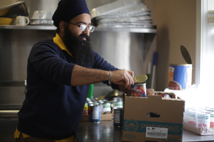 A man with a beard, glasses, and a turban packs canned food into a cardboard box in a kitchen.