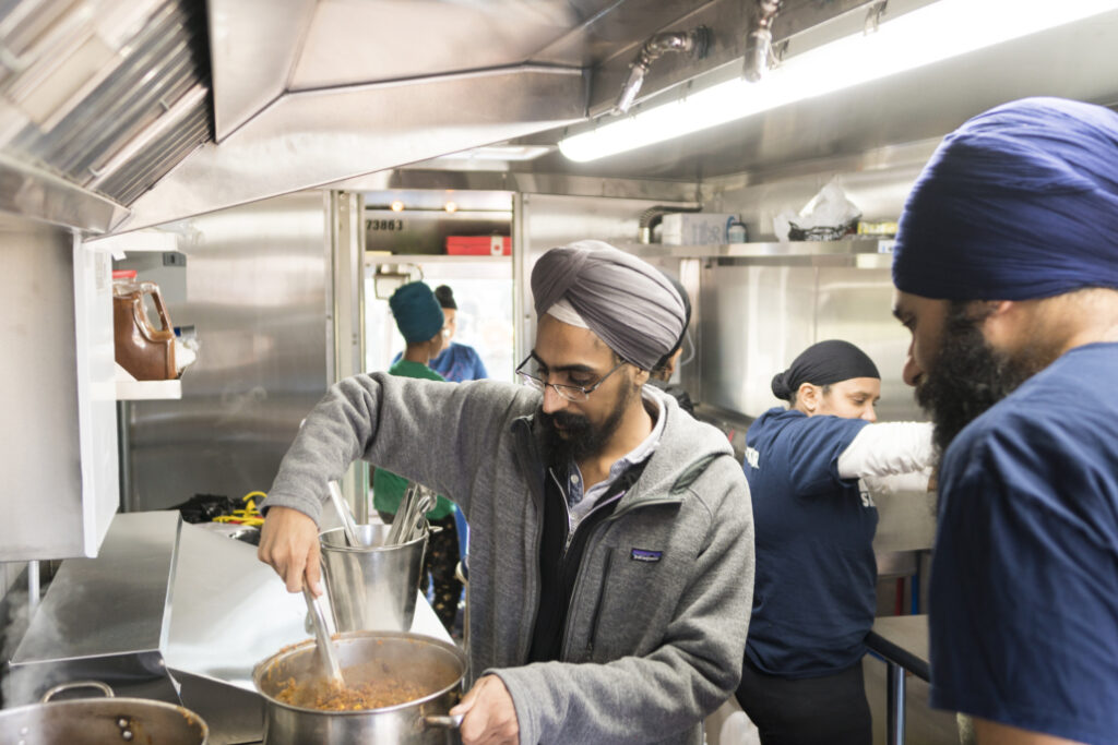 A man wearing a turban and glasses stirs a pot of food in a commercial kitchen.