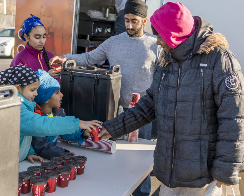 A woman in a pink hat hands a cup to a girl in a blue jacket.