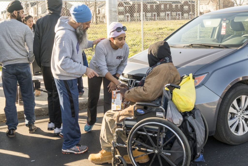 People distribute food to a person in a wheelchair near a gray car in an outdoor setting.