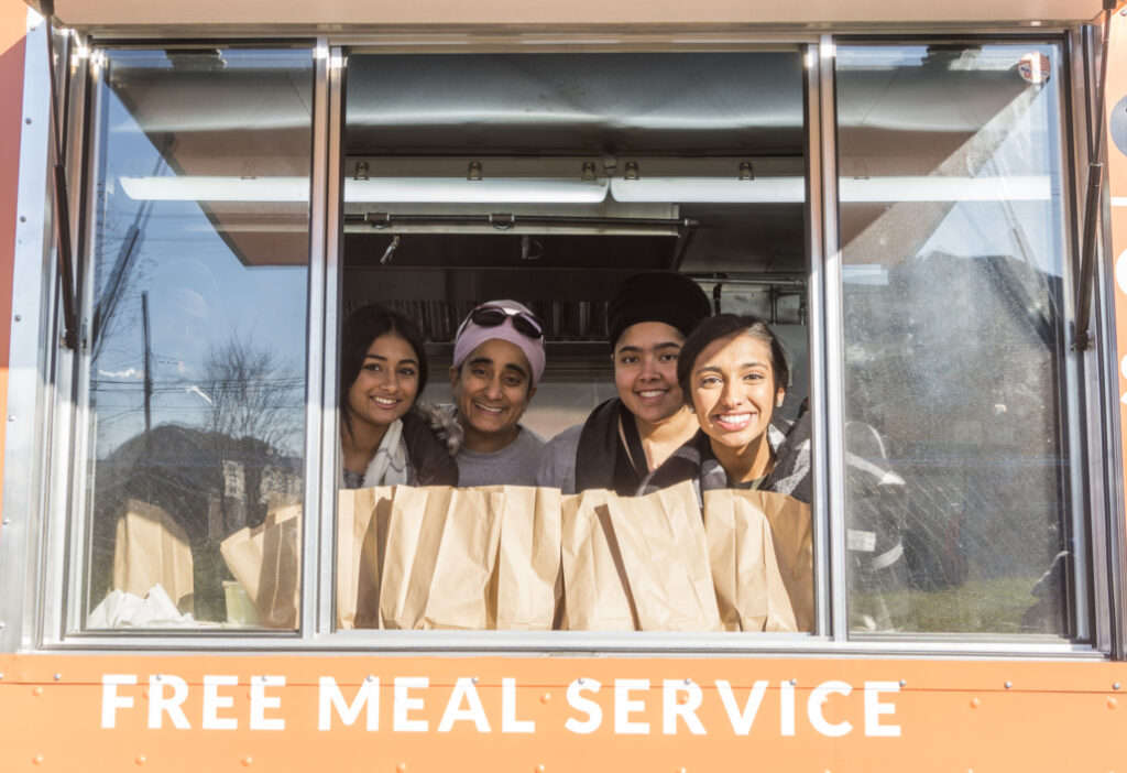 Four smiling women stand inside a food truck with a sign that reads "FREE MEAL SERVICE."
