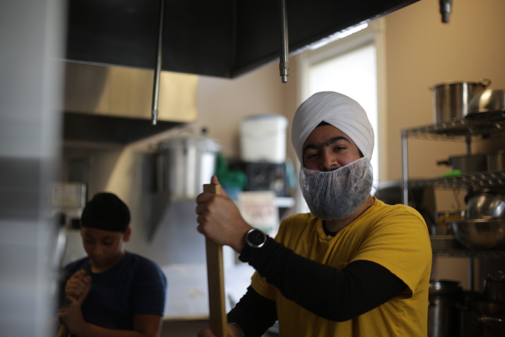 A man wearing a turban and face mask stirs a large pot in a busy kitchen.