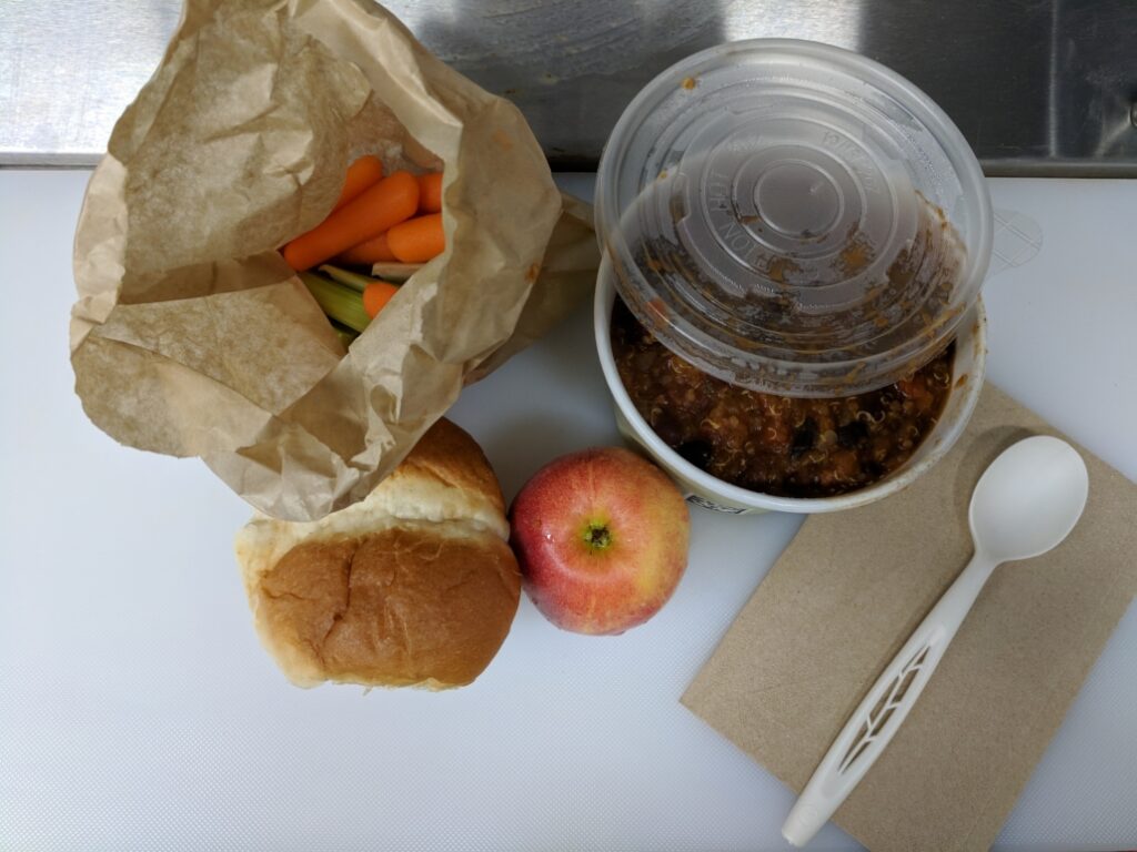 A meal with a bread roll, apple, baby carrots in a paper bag, a cup of chili.