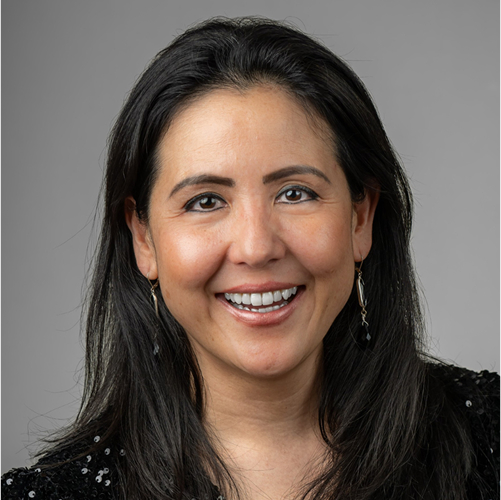 A woman with long dark hair, smiling, wearing earrings and a black top against a gray background.