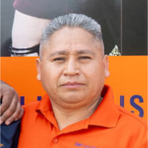 A man with short gray hair wearing an orange polo shirt stands in front of an orange background.
