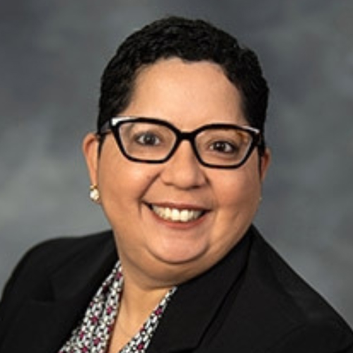 A woman with short dark hair, glasses, and earrings, smiling in a professional black blazer against a gray background.