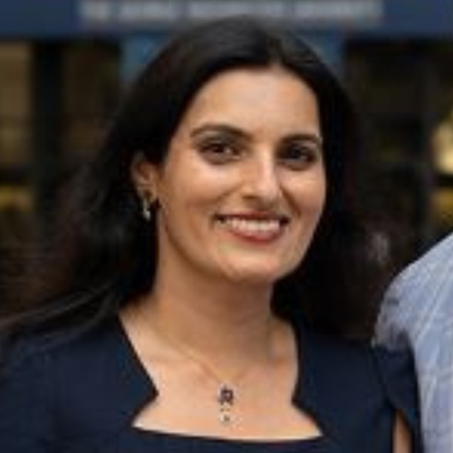 A woman with dark hair, wearing a navy blazer and jewelry, smiling outdoors in front of a blurred background.