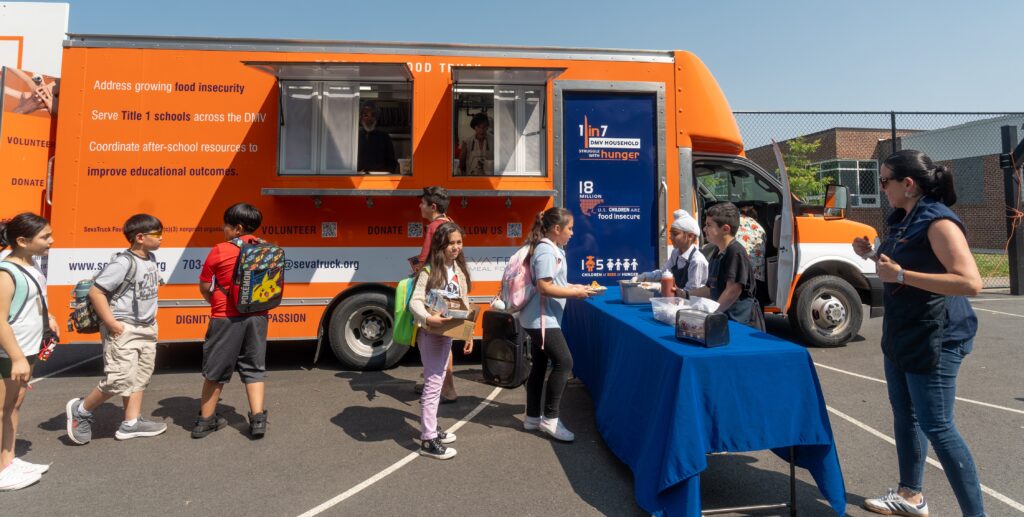 Children line up to receive food from a mobile food truck with orange and blue branding, outdoors on a sunny day.