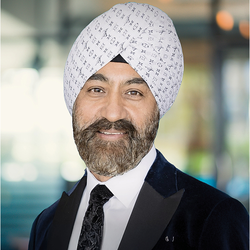 A man with a beard wearing a white turban with black symbols, a tuxedo, and a white shirt, smiling indoors.