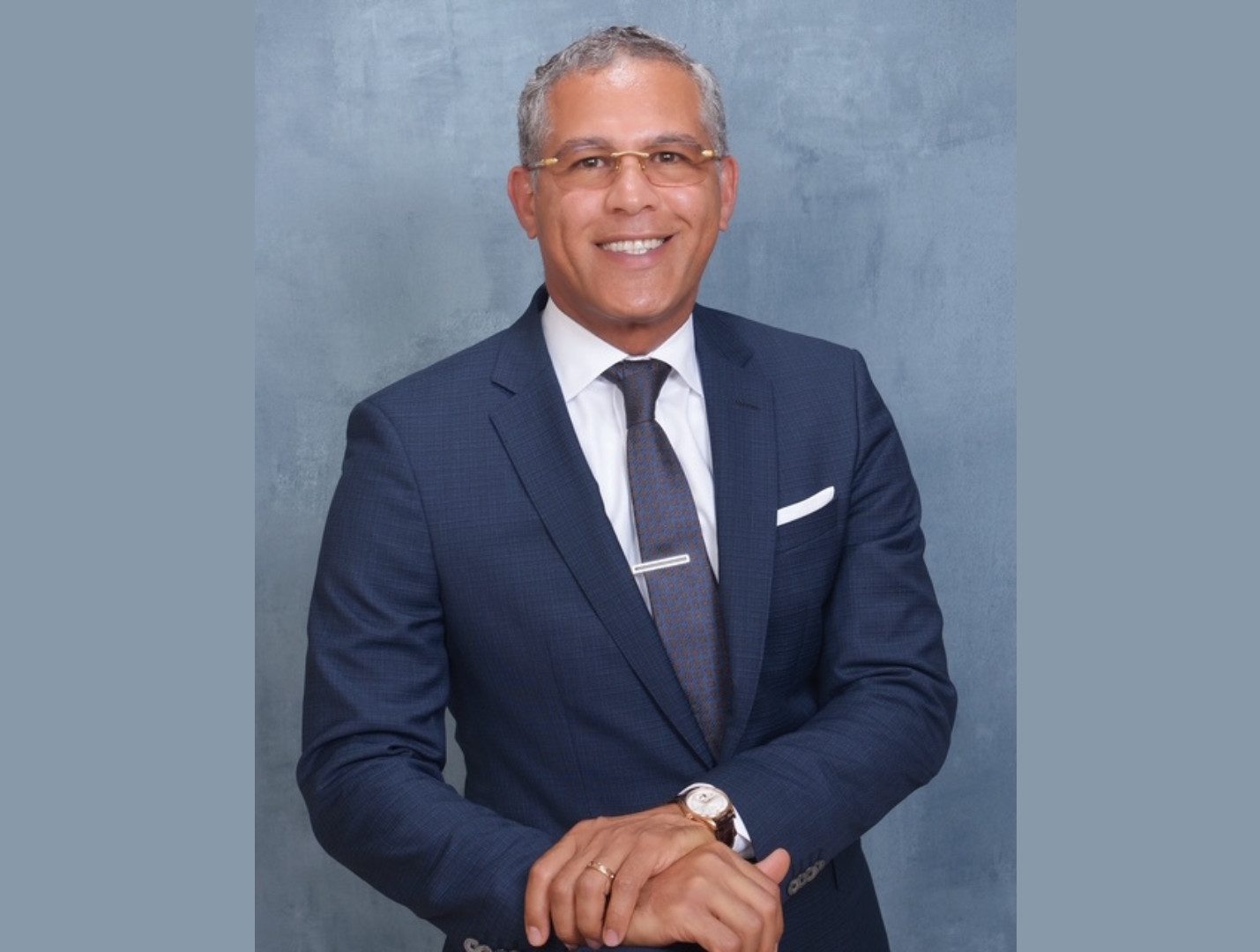 A smiling man in a navy suit, white shirt, and patterned tie