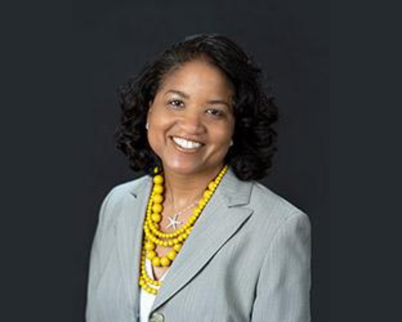A smiling woman with curly black hair wearing a light gray blazer and yellow beaded necklaces against a dark background.