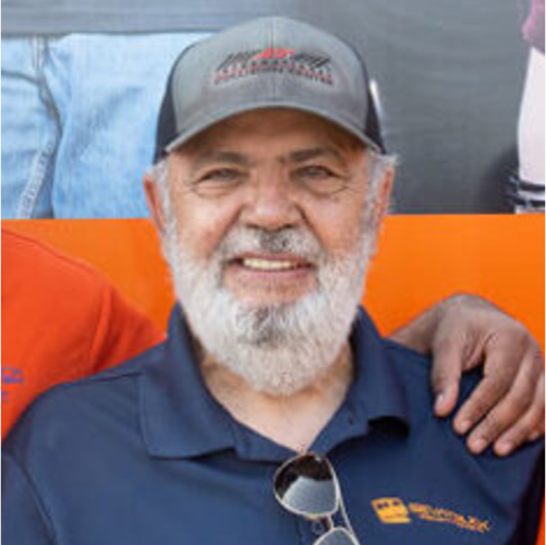 A smiling elderly man with a white beard wearing a gray cap and navy blue polo shirt, standing outdoors with others.